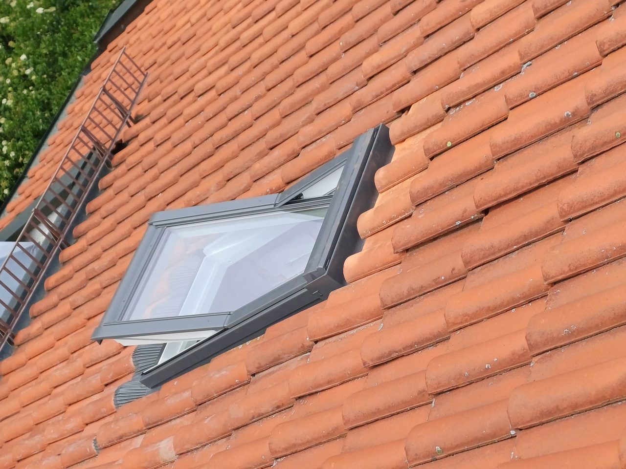 Dachflächenfenster in einem älteren roten Tonziegeldach, mit grauer Verblechung, daneben eine Dachleiter